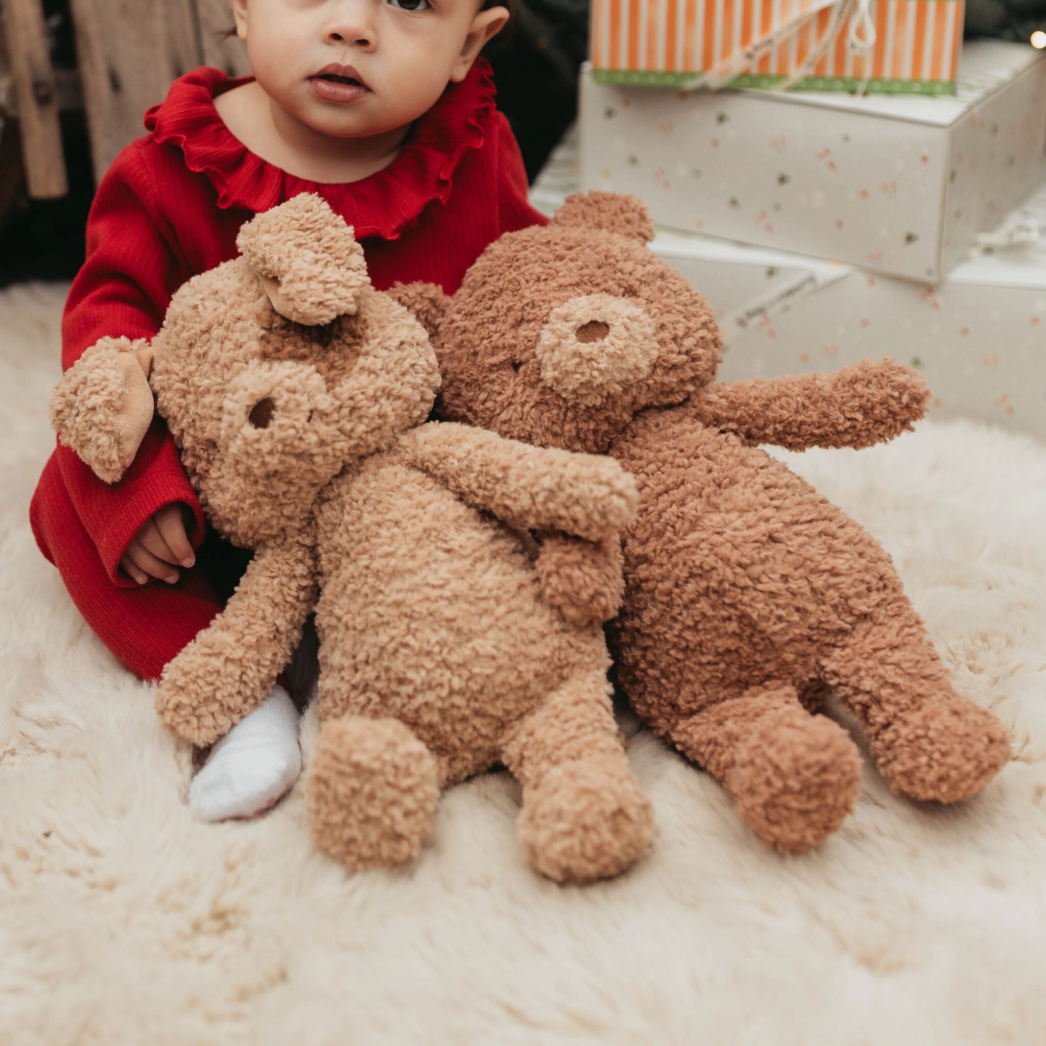 Child in a red outfit holding two brown plush animals on a light-colored surface.