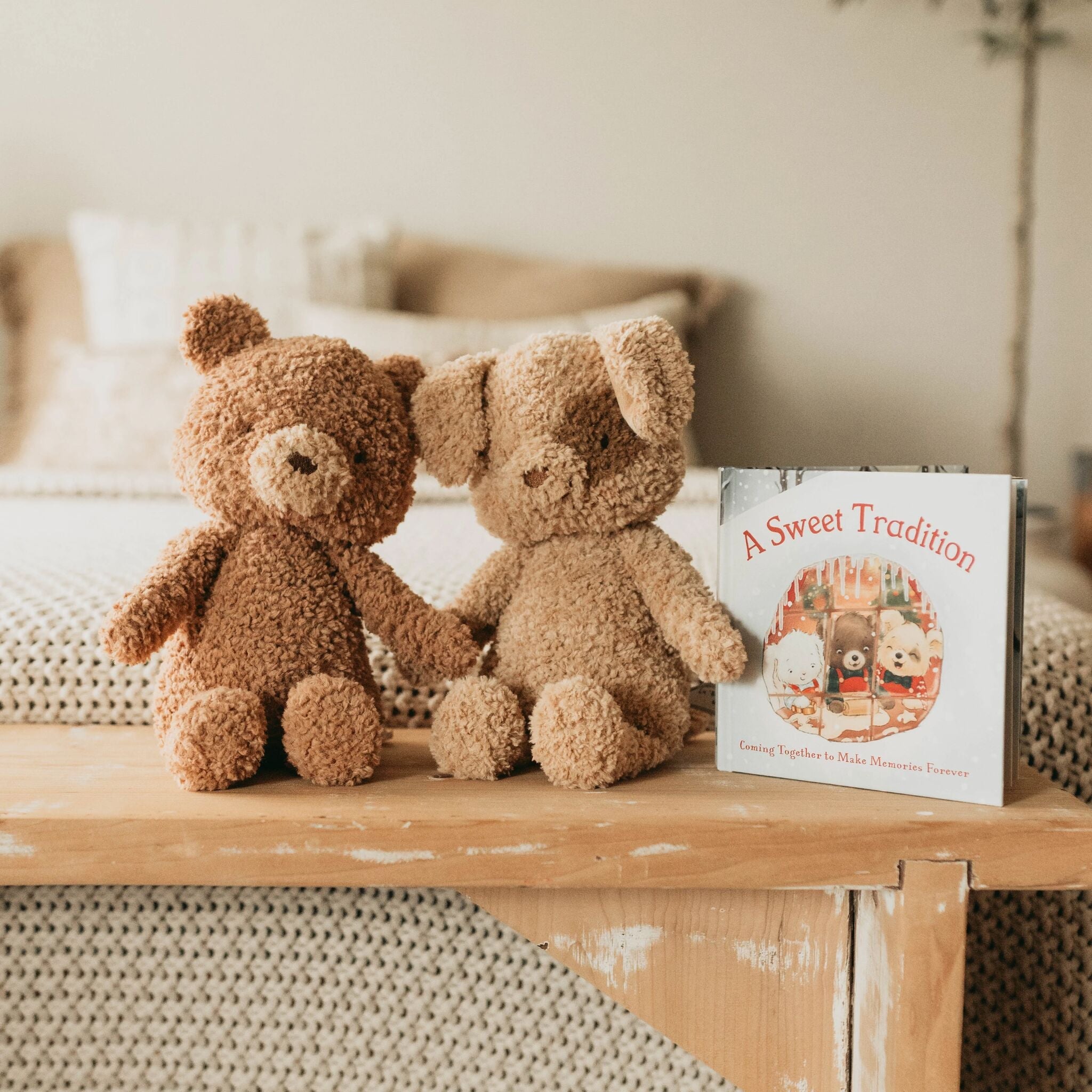 Two brown plush animals holding hands on a wooden surface with a book titled 'A Sweet Tradition' in the background.
