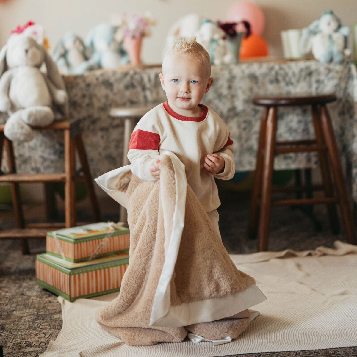 A baby standing on a rug in a cozy room, holding a plush tan blanket with white trim. The baby is wearing a cream long-sleeve top with red accents.