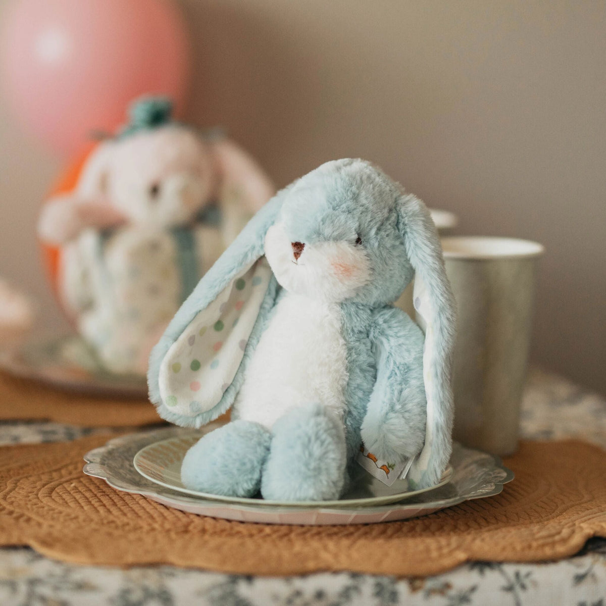 A light blue and white plush bunny toy sitting on a decorative plate on a table, with a blurred second bunny and a pink balloon in the background.