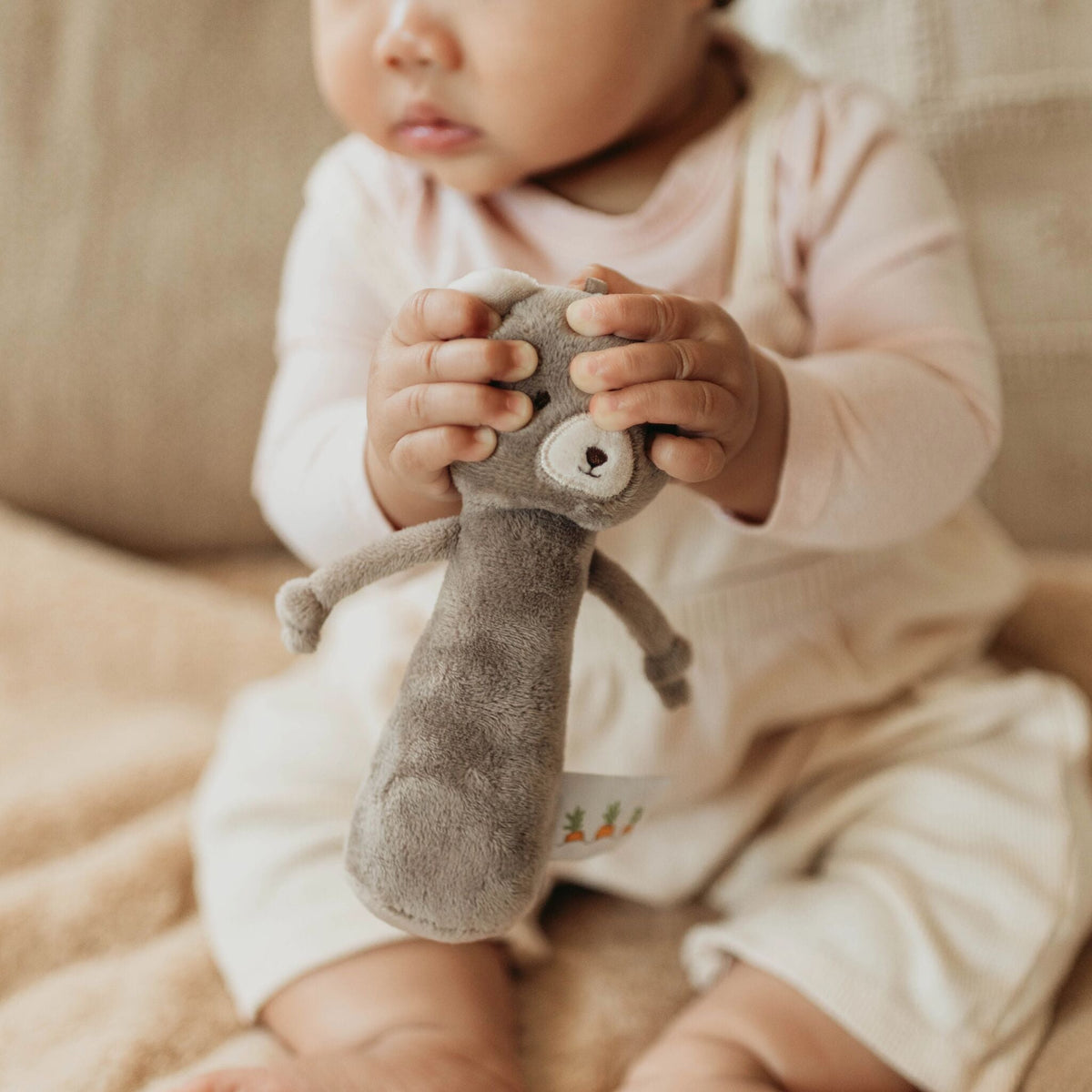 A baby wearing a pale pink ribbed romper sits on a beige blanket, holding a soft brown plush animal toy with a white face.
