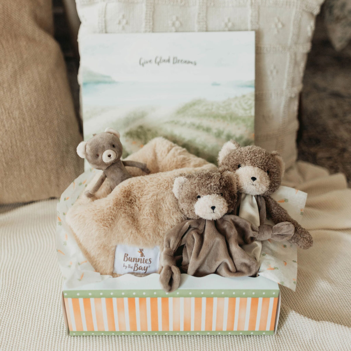 A baby gift set containing three plush teddy bear toys, a soft beige blanket, arranged in a striped gift box on a cozy fabric surface.