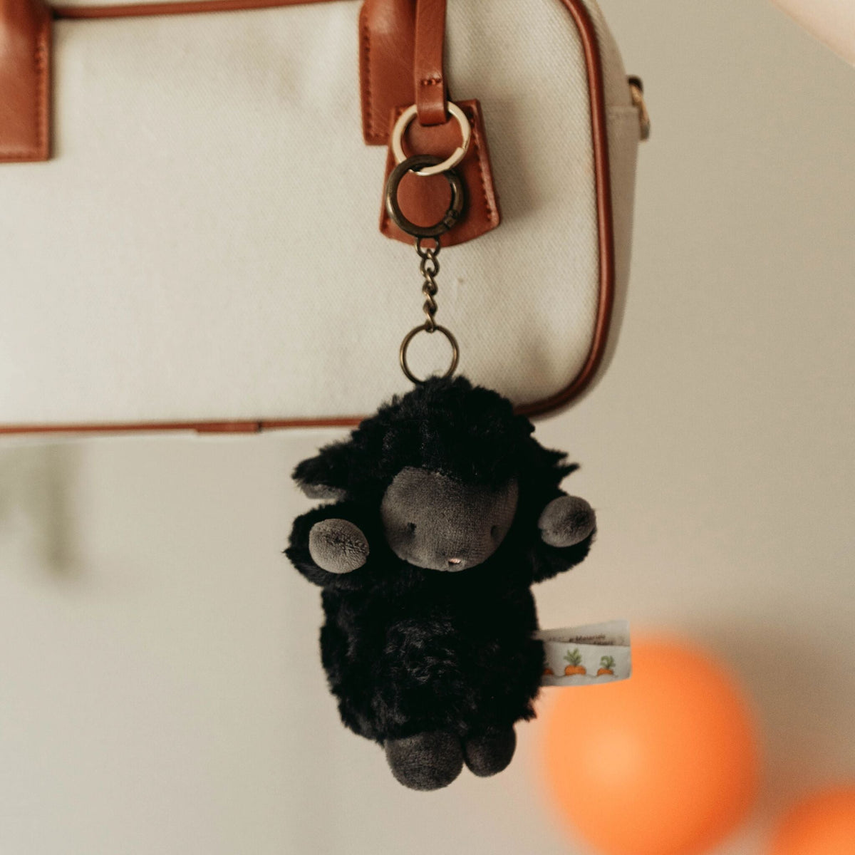 A small black plush sheep keychain with gray details hangs from a cream-colored bag with brown leather trim.