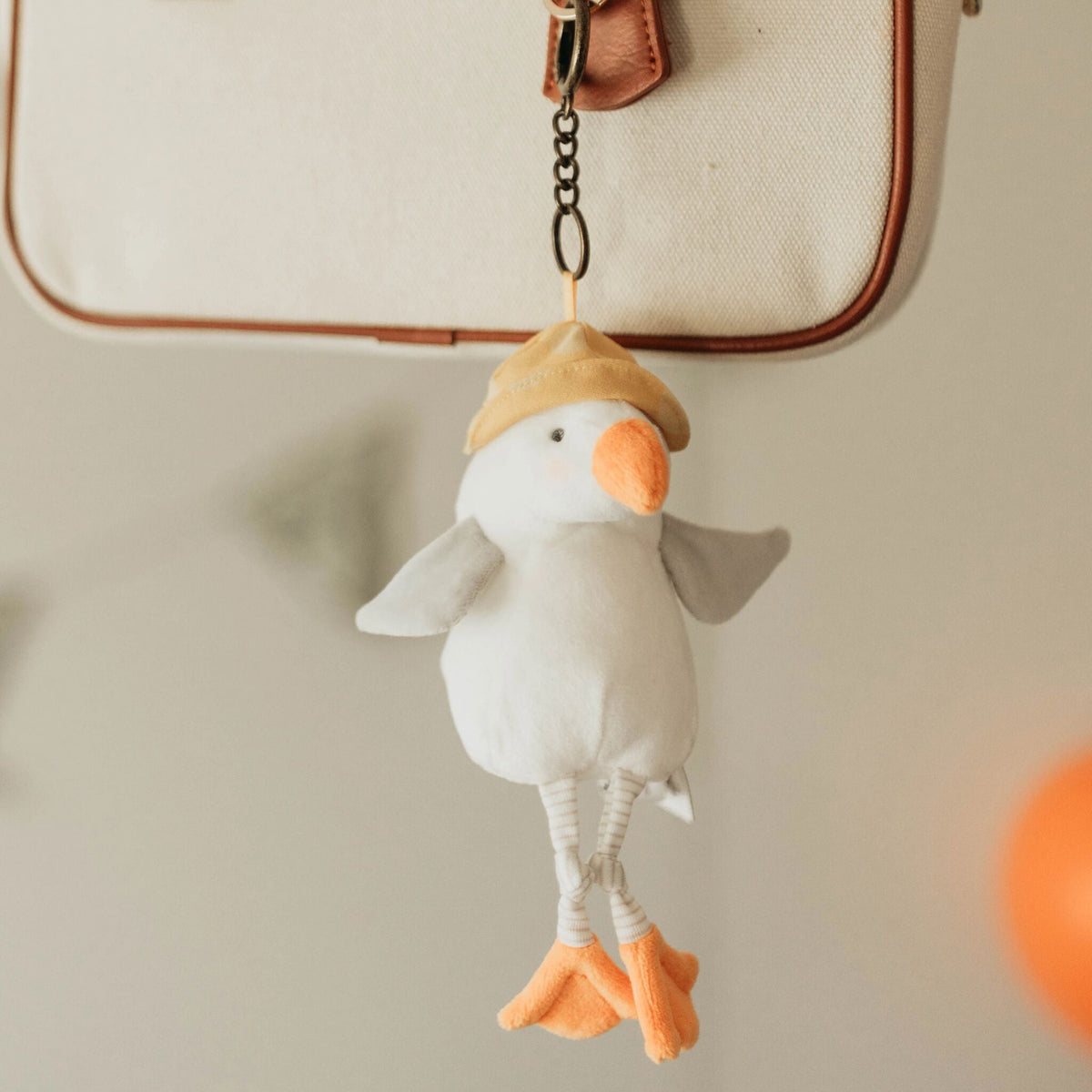 A plush seagull keychain or bag charm with a white body, orange beak and feet, and a tan hat, hanging from a black chain in front of a cream canvas bag with brown trim.