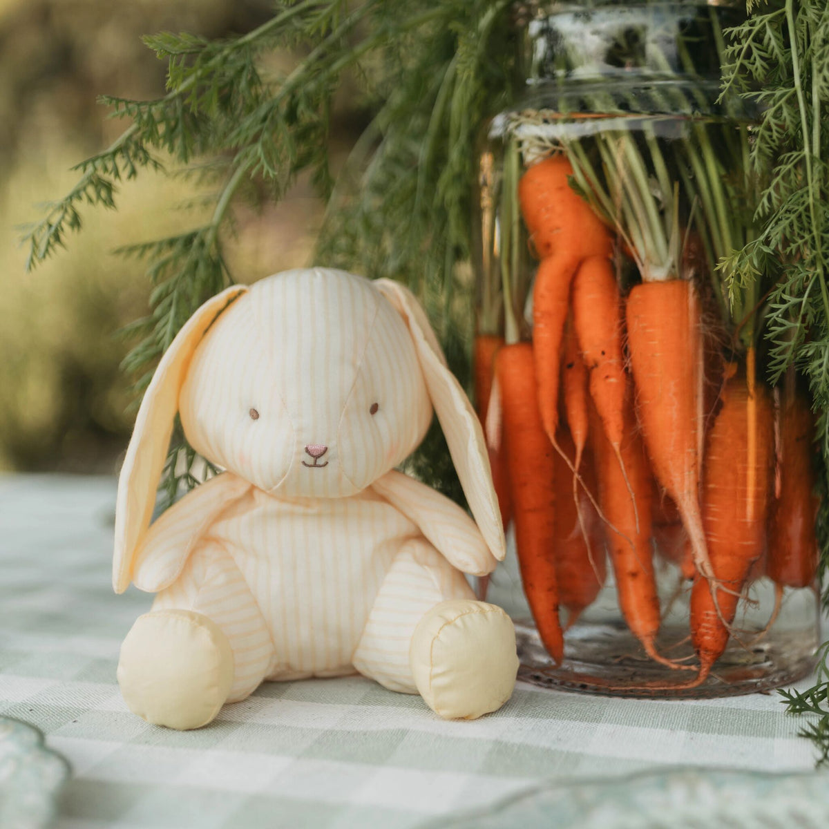 A cream-colored plush bunny doll with a vertically striped fabric body sits on a checkered tablecloth next to a glass jar filled with fresh orange carrots and green tops.