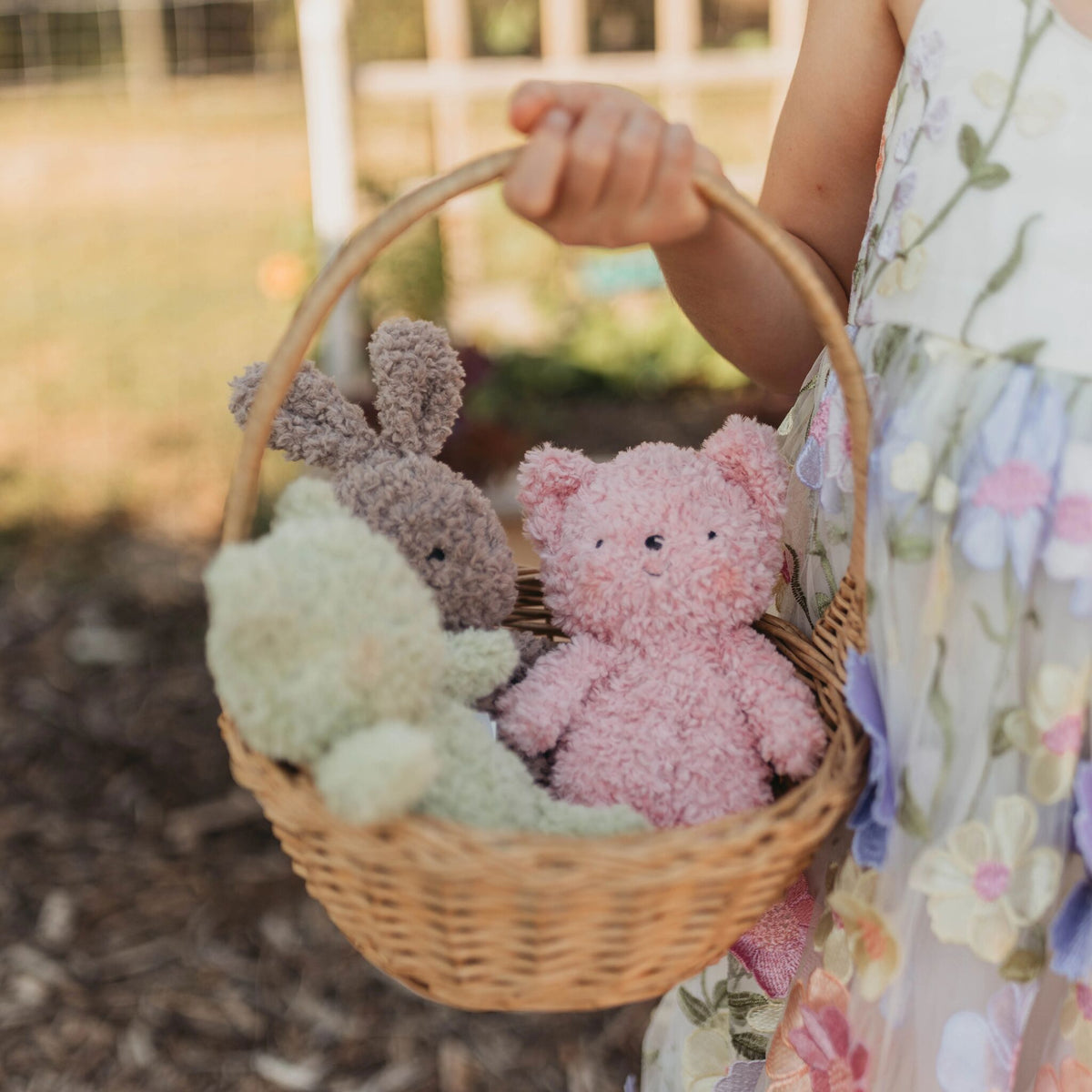 A child in a floral dress holds a wicker basket containing three fuzzy plush toys: a pink cat, a gray bunny, and a pale green frog, photographed outdoors with a blurred natural background.