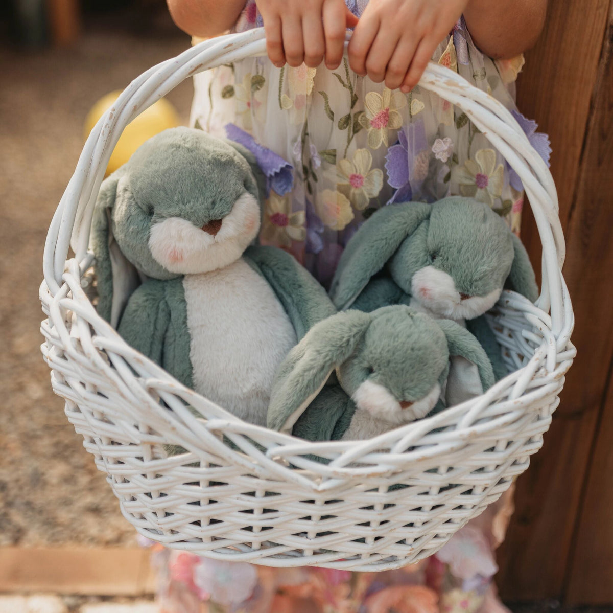 A white woven wicker basket held by a child, containing three plush green bunny toys with white faces.