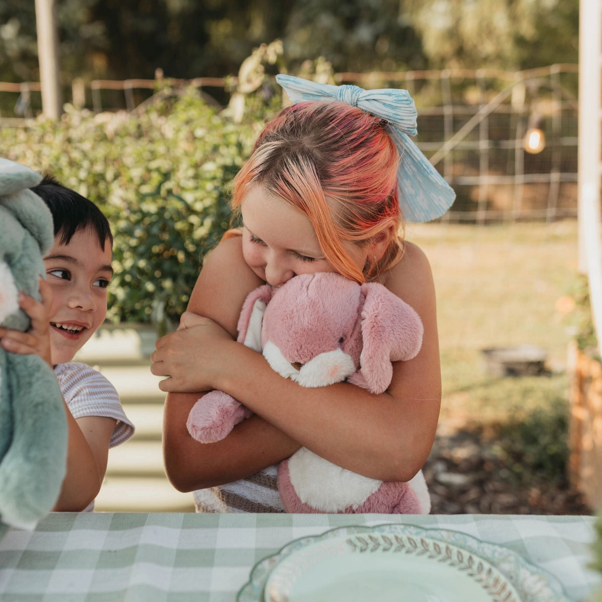 A lifestyle scene featuring a young girl with pink and orange hair holding a pink plush bunny, with a boy partially visible on the left holding a green plush toy. They are seated at a table with a green and white checkered tablecloth in an outdoor garden setting.