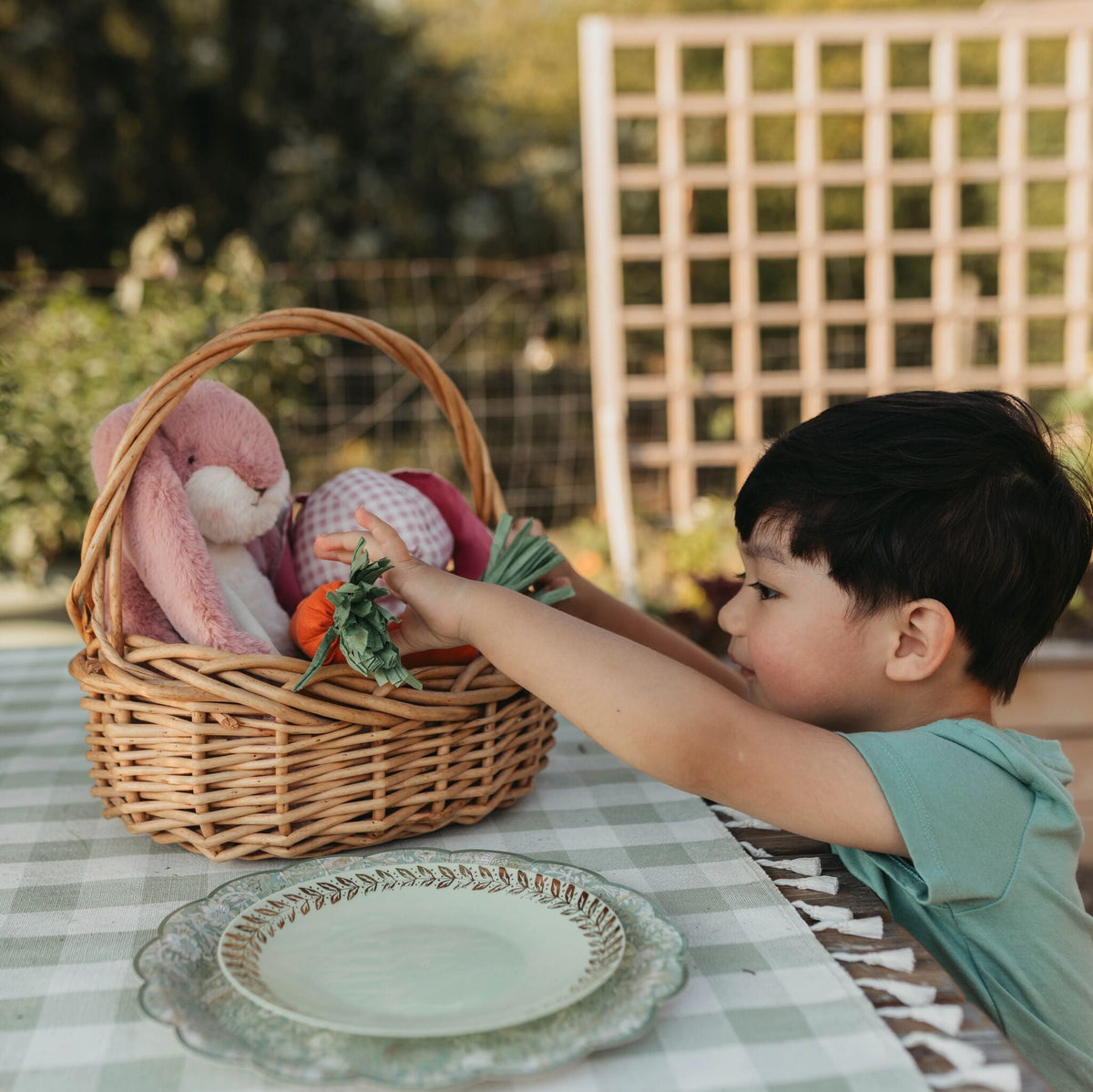 A young boy in a mint green shirt leans over a wicker basket filled with plush toys, including a pink bunny and an orange carrot, set on a green-and-white checkered table outdoors.