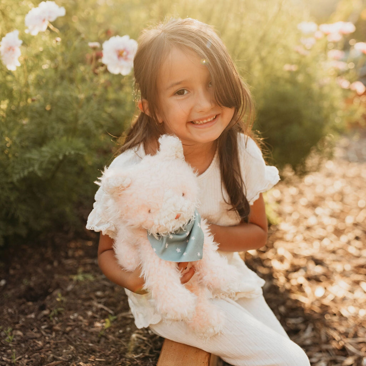 A young girl with long brown hair sits outdoors in a garden, smiling while holding a fluffy pink stuffed cat with a light blue bandana.