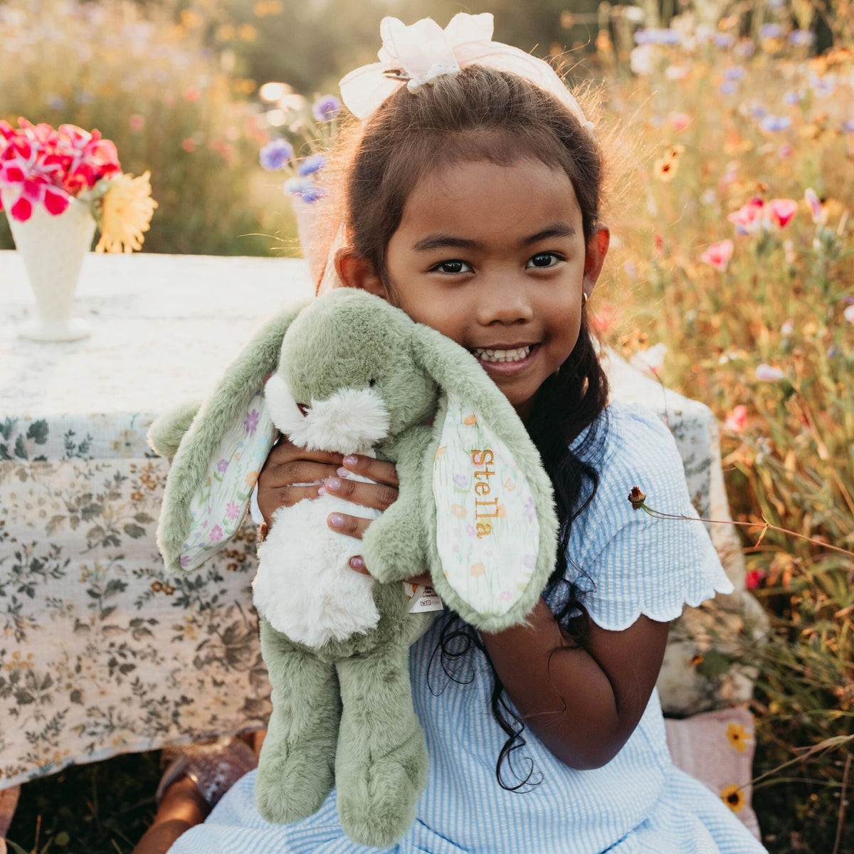 A young girl with a white bow in her hair smiles while holding a personalized plush bunny toy. The bunny is light green with a white belly and features the name 'Stella' embroidered on its ear.