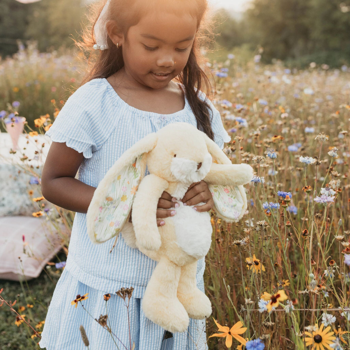 A young girl in a light blue dress holds a yellow-colored plush bunny with pastel floral ears in a sunlit wildflower field.