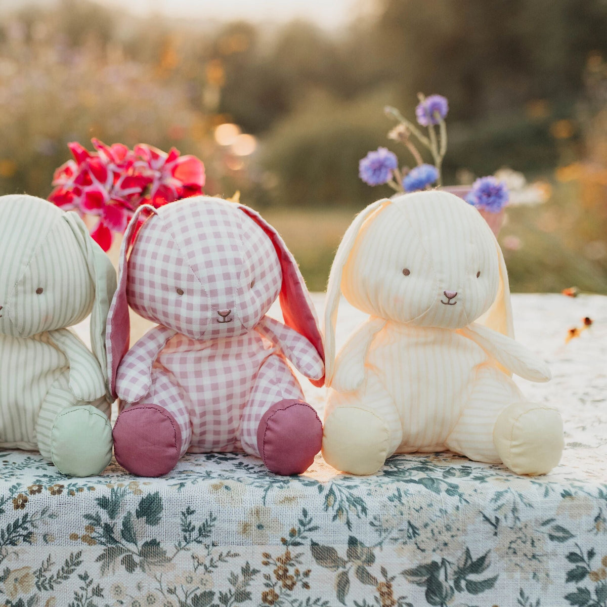 Three handmade plush bunny toys in soft fabrics are arranged on a floral tablecloth outdoors. The left bunny is cream and green, the center bunny wears a pink gingham outfit with pink feet, and the right bunny is cream with pale yellow feet.