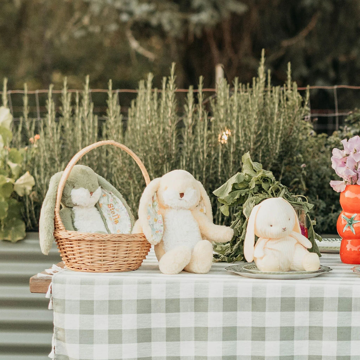 Three plush bunny toys arranged on a table outdoors, with one in a wicker basket, one sitting on the table, and one sitting on a plate. The scene includes a green-and-white checkered tablecloth, leafy greens, and a blurred garden background.