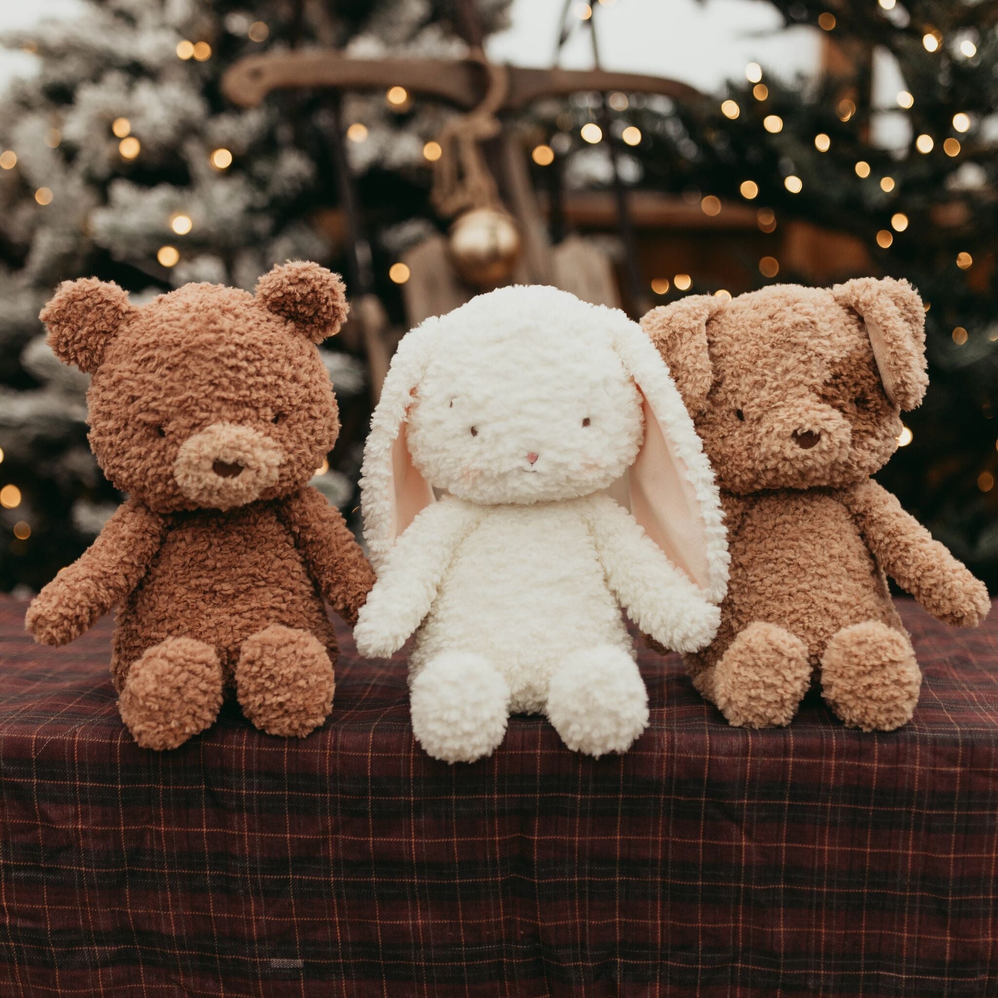 Two brown teddy bears and a white rabbit plush toy on a checkered surface with a blurred festive background.