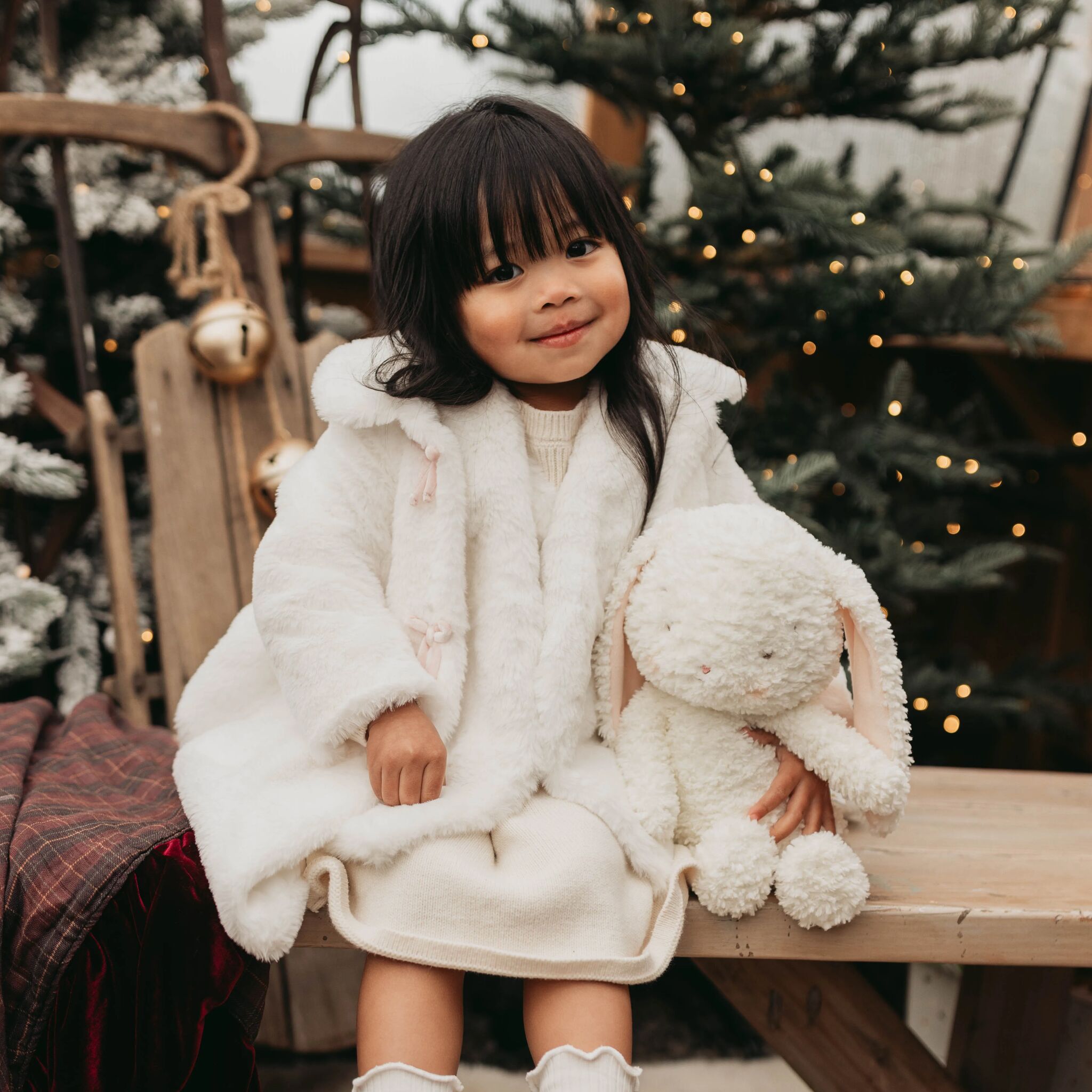 Child in a white fluffy coat holding a plush toy in front of a decorated Christmas tree.