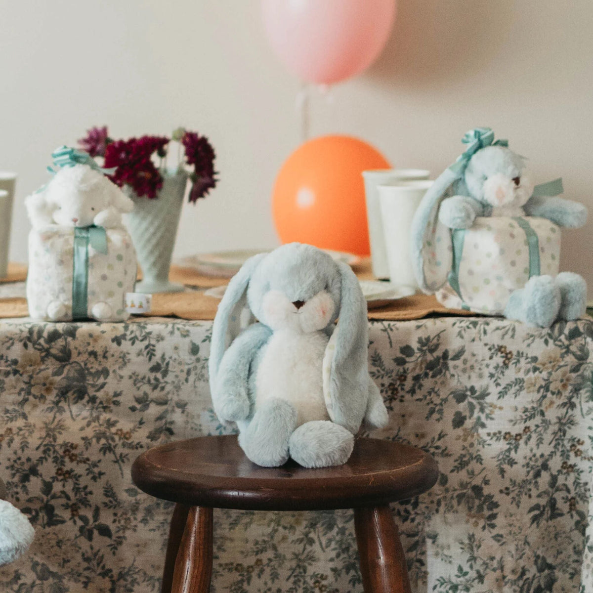 A plush light blue and white bunny toy sits on a small wooden stool in a styled indoor setting with a floral tablecloth, gift boxes, flowers, balloons, and cups in the background.