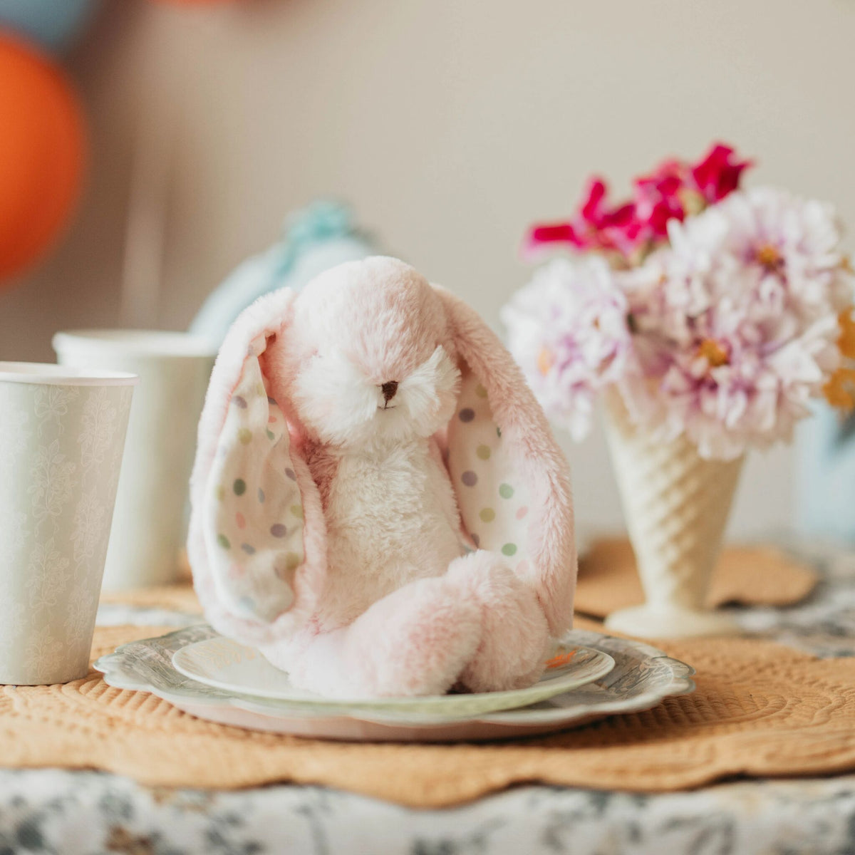 A soft pink plush bunny toy with long floppy ears sits on a small plate on a table. The scene is styled with a cup, a vase of flowers, and a woven placemat in a warm indoor setting.
