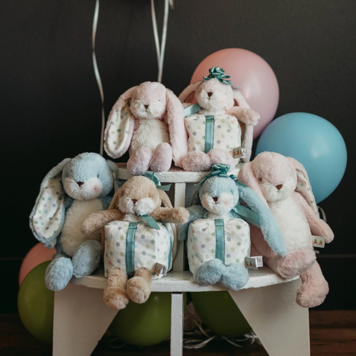 Five pastel plush bunny toys arranged on a small white pedestal. Colorful balloons in pink, blue, green, and peach are behind the display against a dark background.