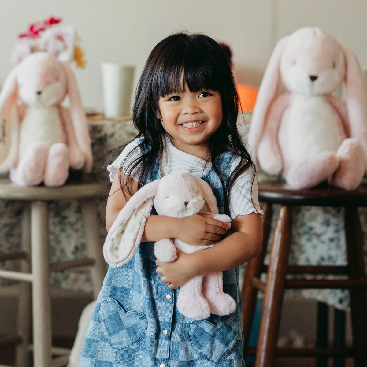 A young girl with dark hair smiles while holding a light pink plush bunny. She is wearing a blue and white checkered pinafore dress over a white t-shirt.
