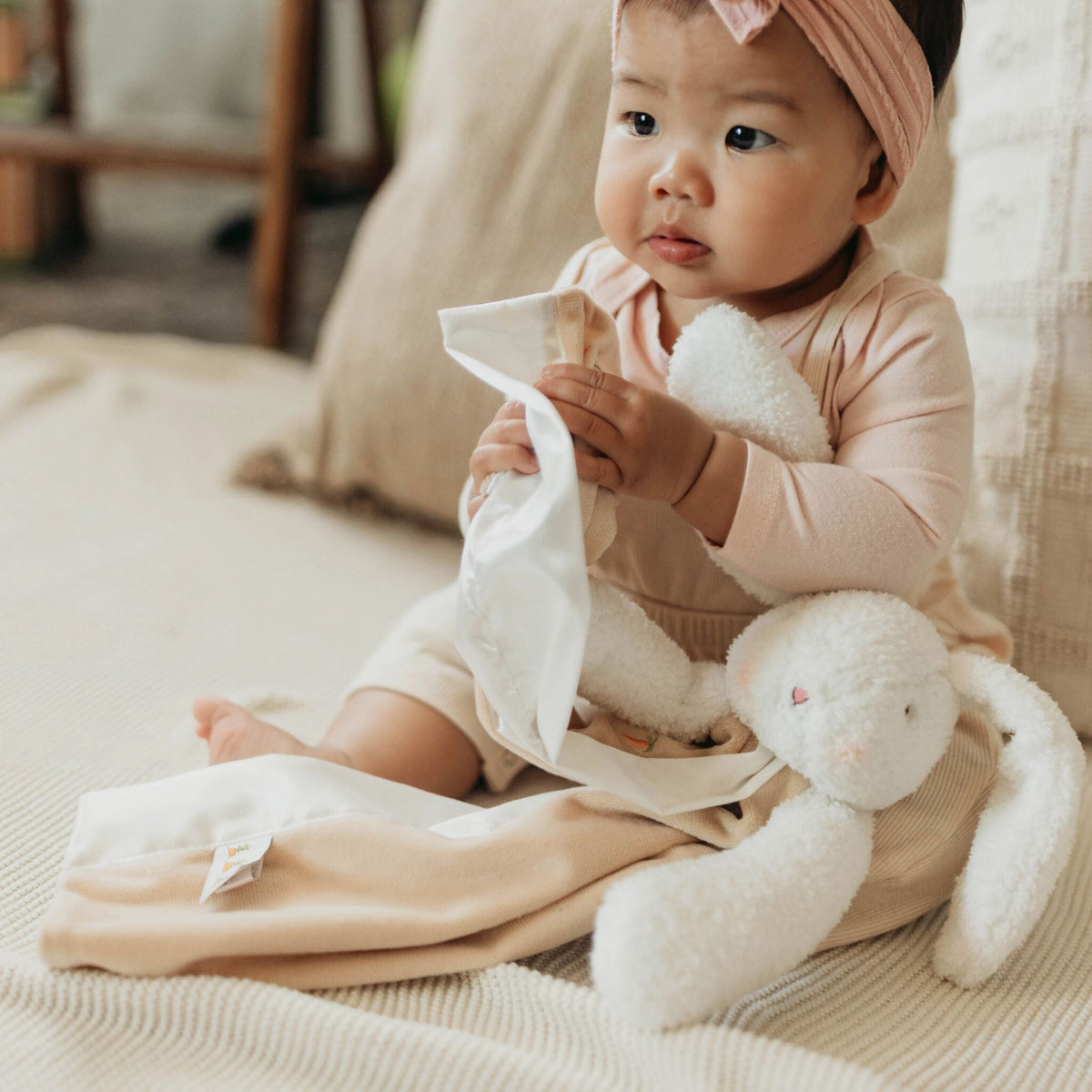 A baby sitting on a cream-colored blanket, wearing a light pink long-sleeve romper and a pink bow headband, holding a white fabric toy or cloth next to a white plush bunny.