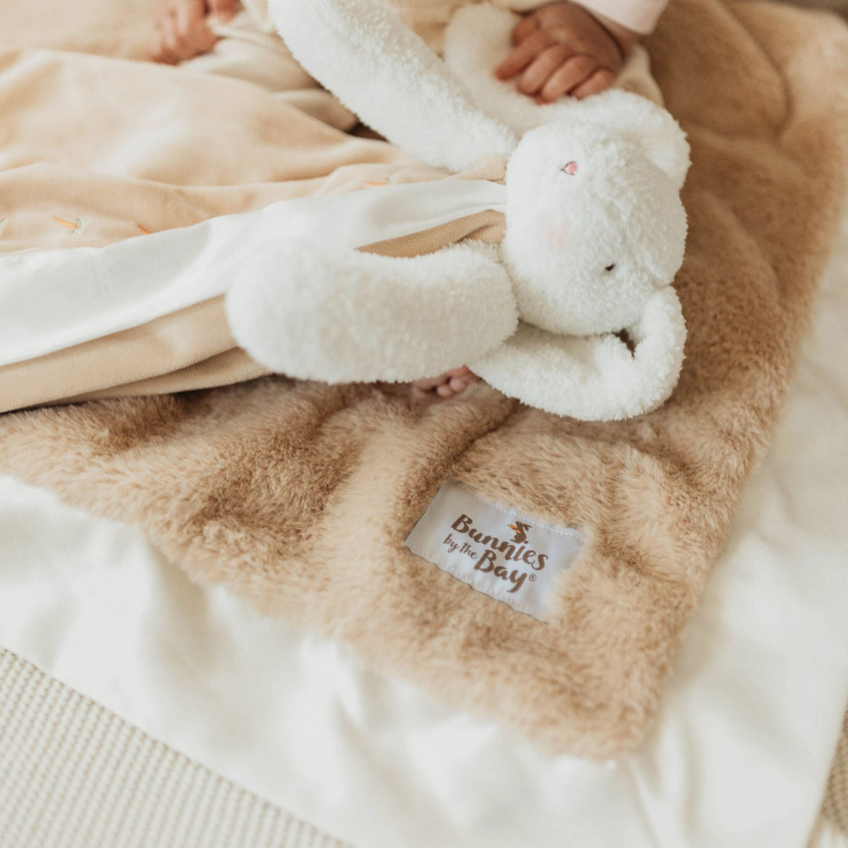 A close-up of a soft plush blanket in warm beige with a white fabric sheet underneath, featuring a small white label that reads 'Bunnies by the Bay' and a white stuffed bunny held by a baby's hand.