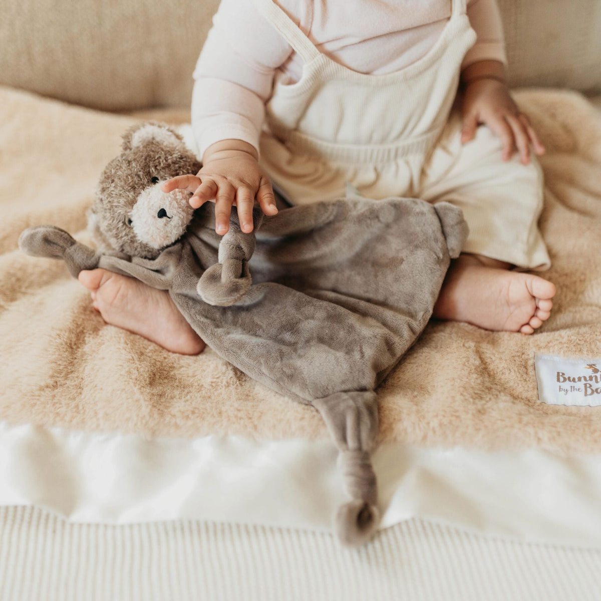 A baby sitting on a beige blanket, holding a soft plush bear comforter with a knotted arms and legs. The baby is wearing cream-colored overalls and a pale pink long-sleeve top.