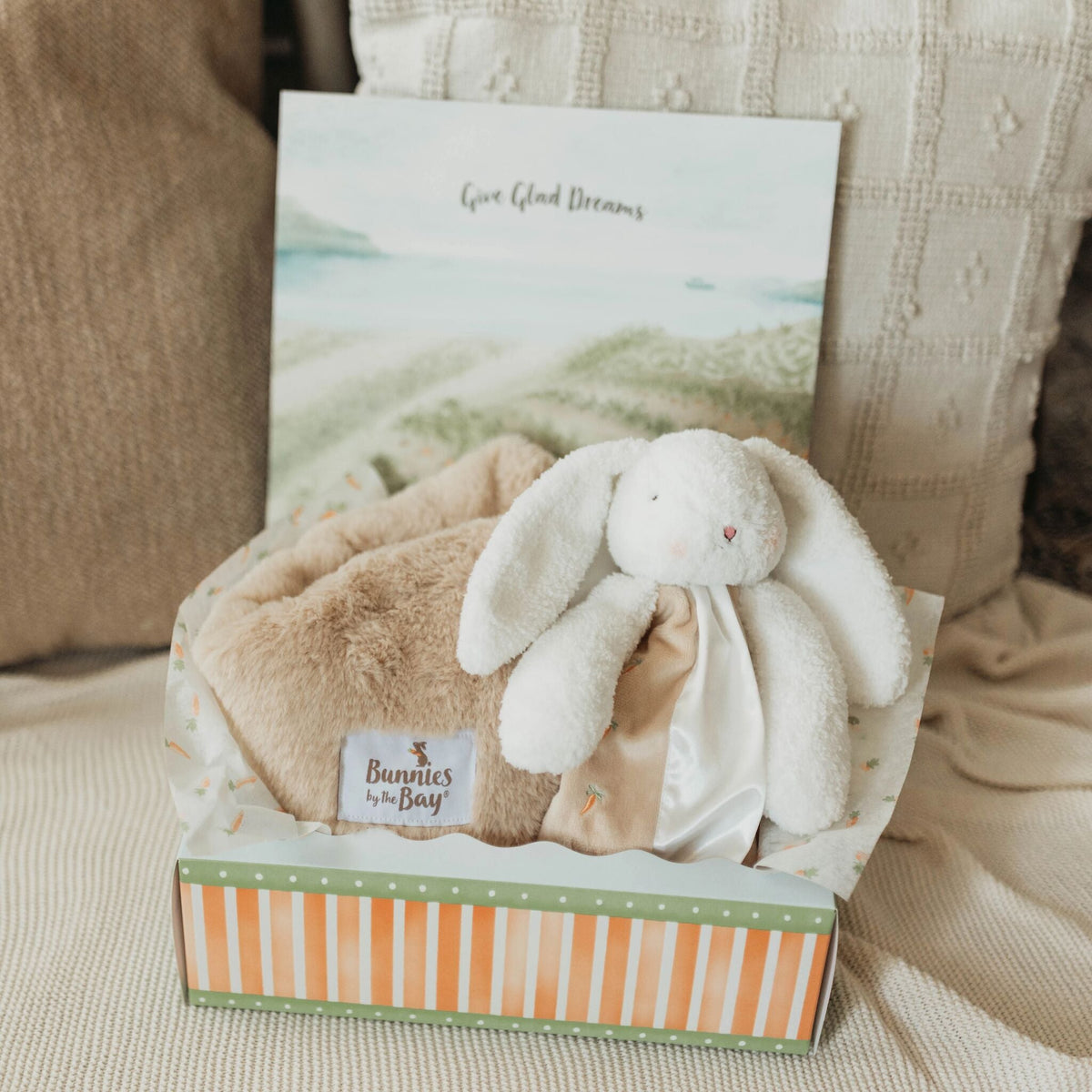 A baby gift set in a striped box featuring a white plush bunny snuggler, a beige blanket, styled on a soft beige fabric surface with textured pillows in the background.