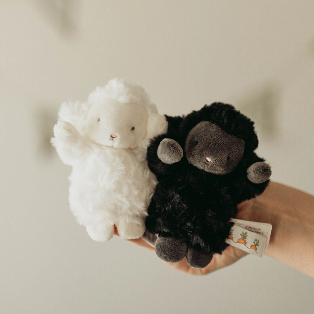 Two plush sheep toys, one white and one black, held in a person's hand against a soft beige background.