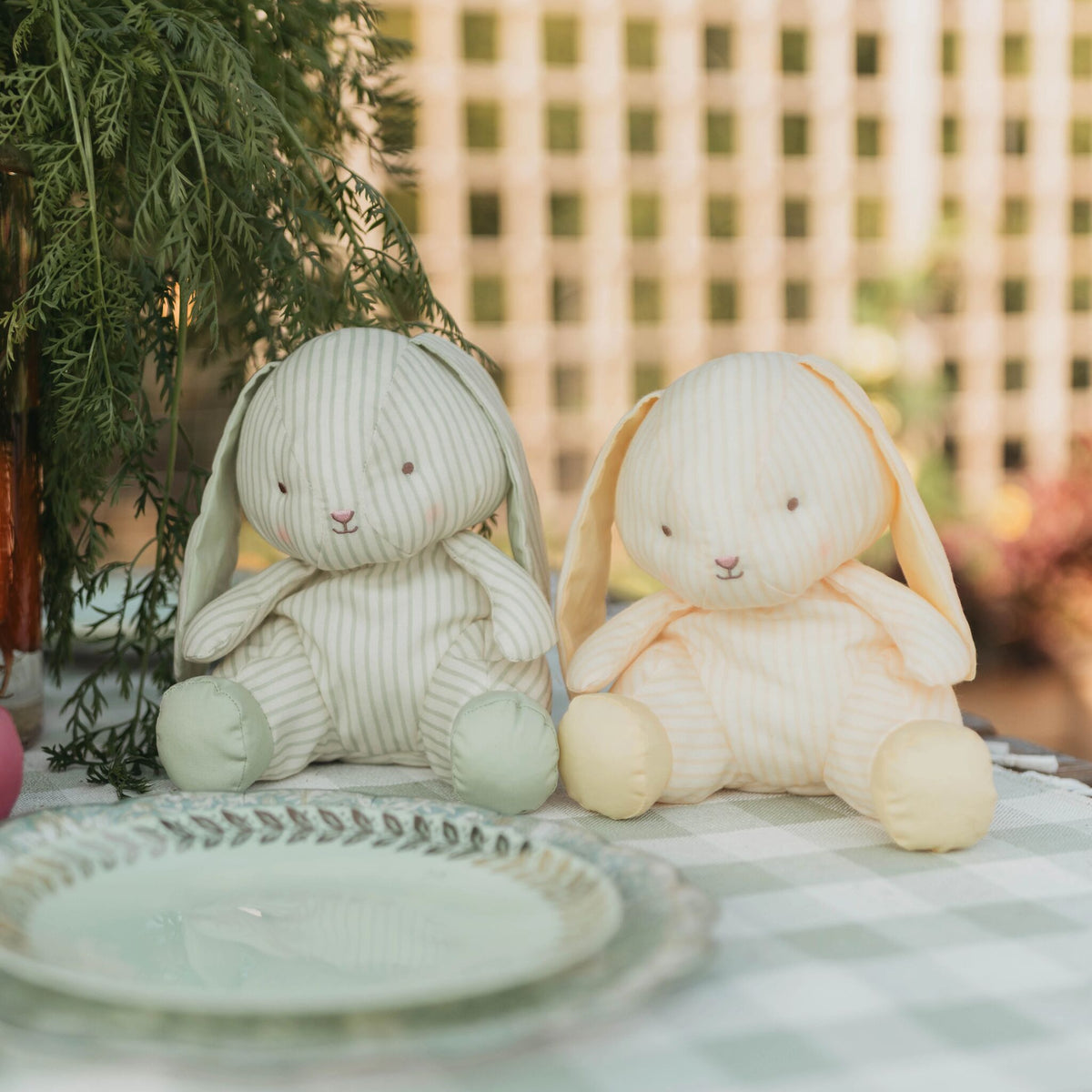 Two plush bunny toys with long floppy ears sit on a checkered tablecloth outdoors. One bunny is light sage green and cream striped, and the other is cream and pale yellow striped.