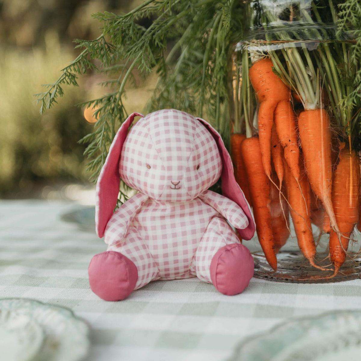 A pink and white gingham stuffed bunny plush toy with pink ears and feet sits on a checkered tablecloth next to a glass jar filled with orange carrots and green tops.