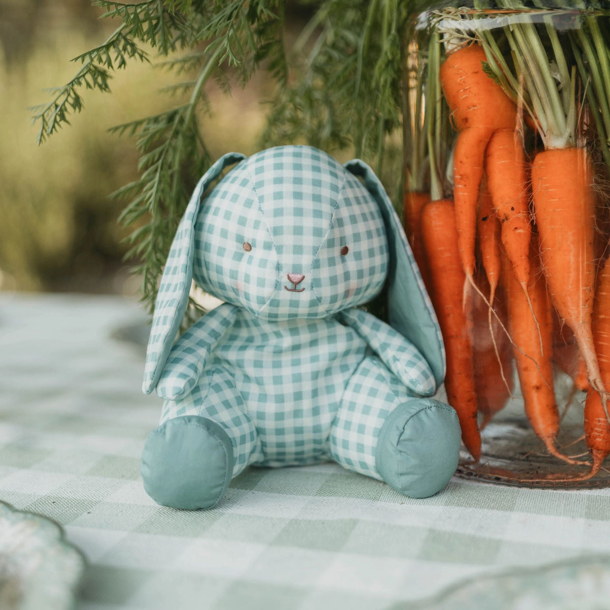 A plush bunny doll in a light blue and white gingham pattern sits on a matching checkered tablecloth next to a glass jar filled with freshly harvested orange carrots and green foliage.