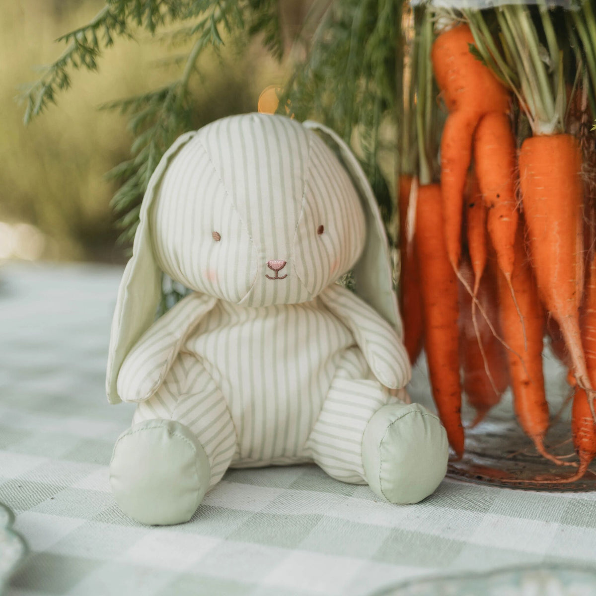 A handmade plush bunny doll with cream and white vertical stripes, sitting on a green and white checkered tablecloth next to a rustic glass jar filled with fresh orange carrots and greenery.