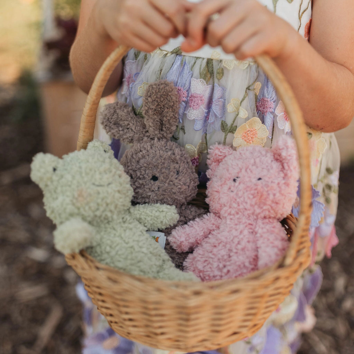 A wicker basket held by a child wearing a floral dress, containing three fuzzy plush animals in pastel green, brown, and pink.