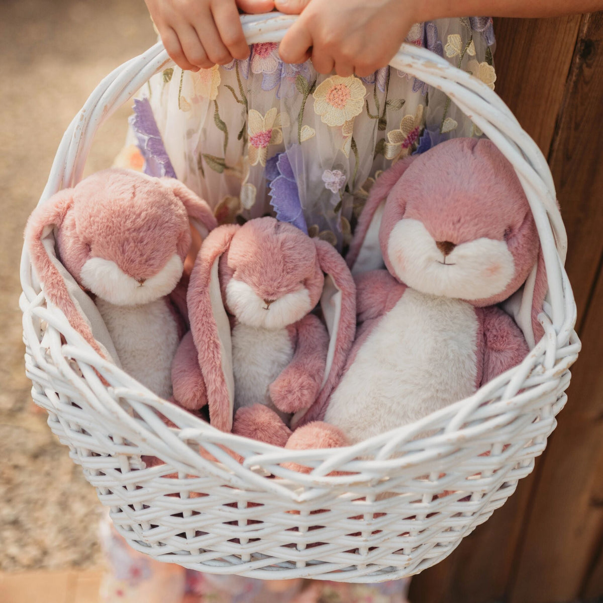 Three pink and white plush bunny toys nestled inside a white wicker basket held by a child's hands, with a floral dress visible in the background.