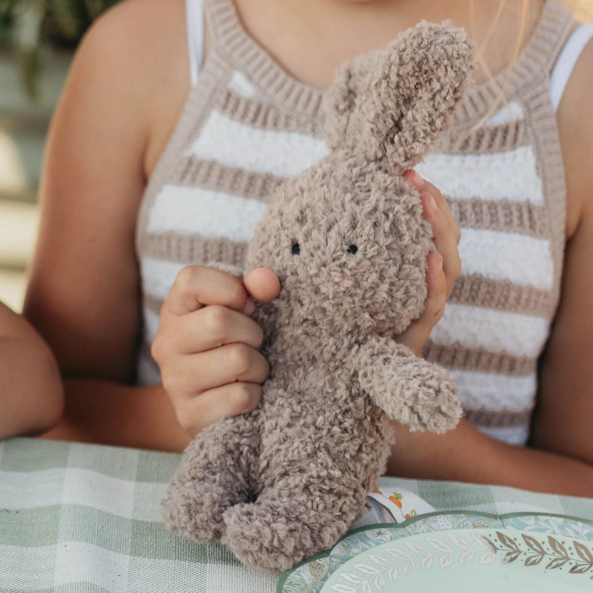 A child in a beige and white striped knit top holds a small, curly-fuzzy brown plush bunny toy at a table with a green and white checkered tablecloth.