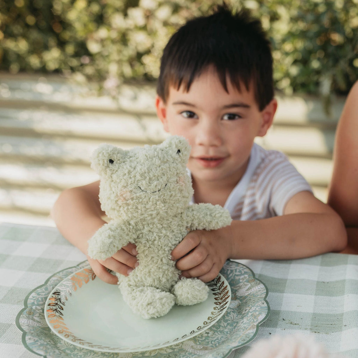A young boy sitting at a table outdoors holds a cream-colored, textured plush frog on a decorative plate. The background shows a softly blurred garden setting with warm sunlight.