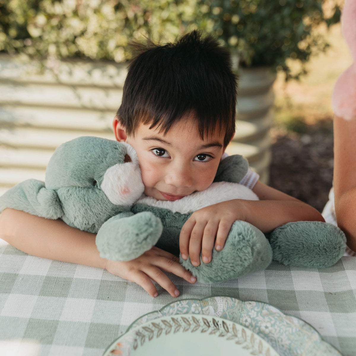 A young boy with dark hair sits at a table outdoors, holding a large mint green plush stuffed animal. The scene is set on a green and white checkered tablecloth with a decorative plate in the foreground and a blurred garden background.
