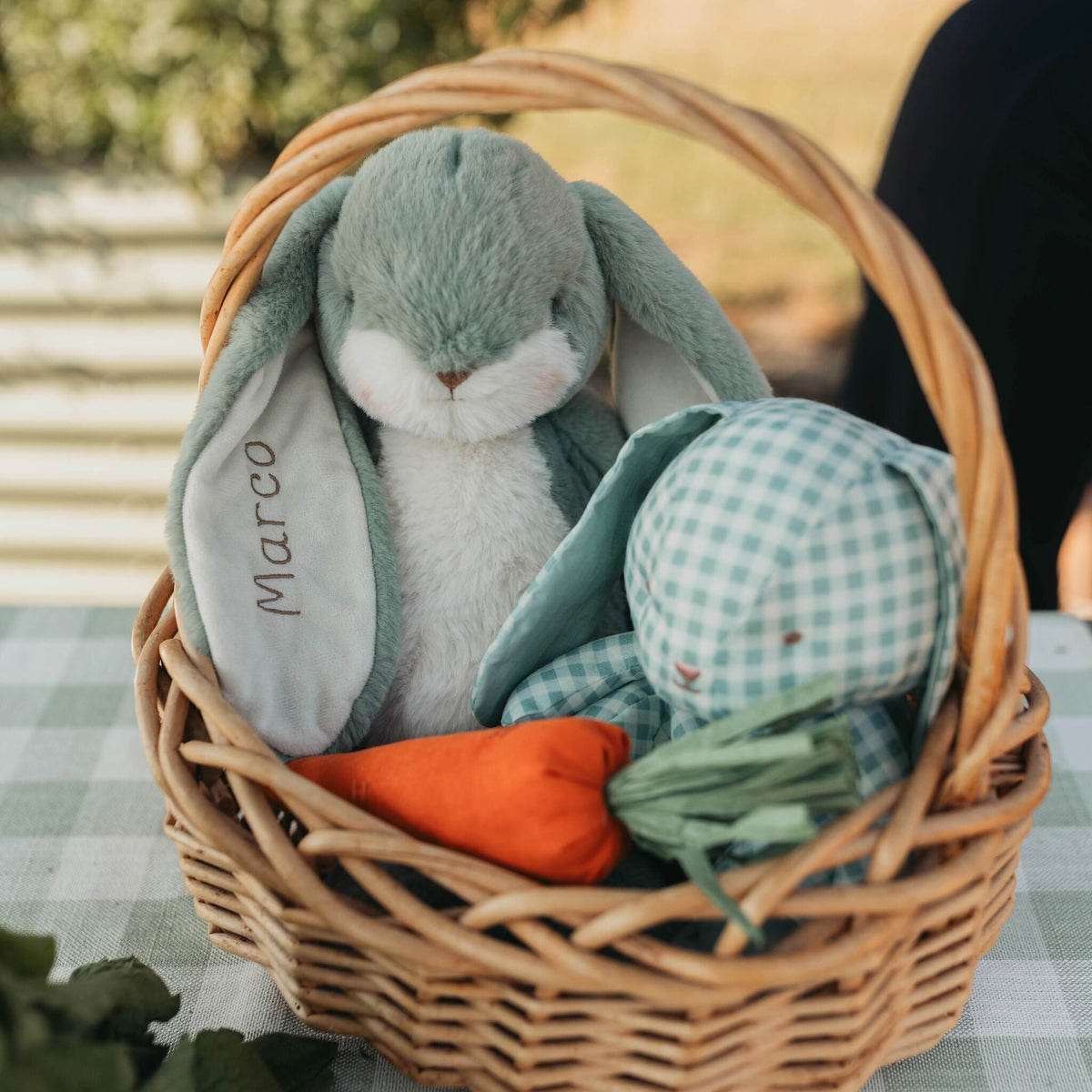 A wicker basket filled with a plush gray-green and white bunny toy, a small orange carrot, and a mint green gingham stuffed animal, set on a checkered tablecloth outdoors.