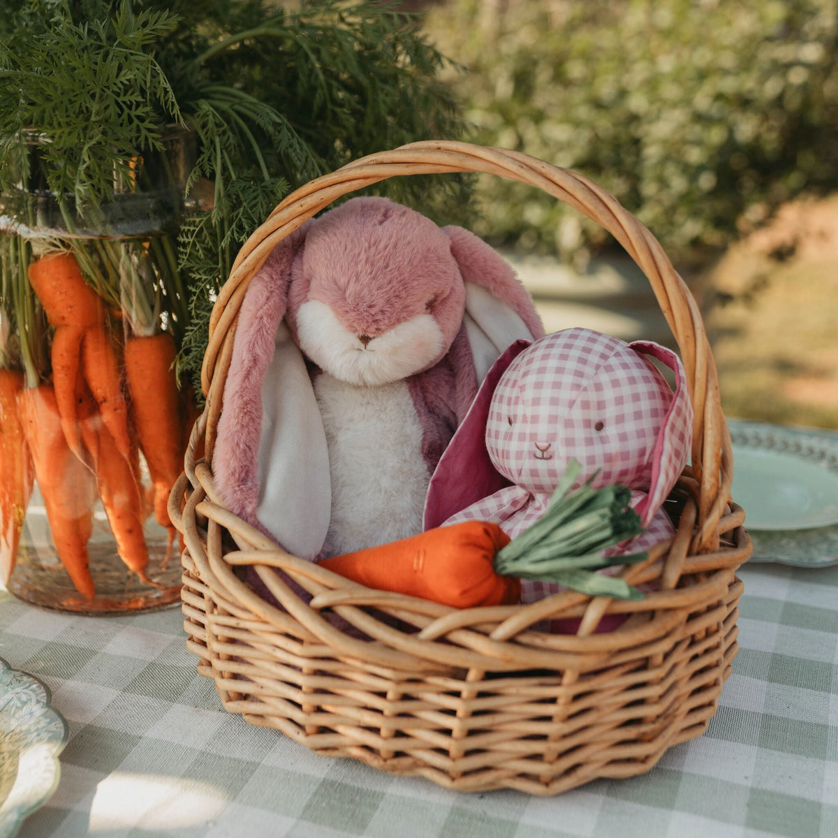 A wicker basket filled with plush bunny toys and toy carrots, placed on a checkered tablecloth outdoors with a glass vase of carrots in the background.