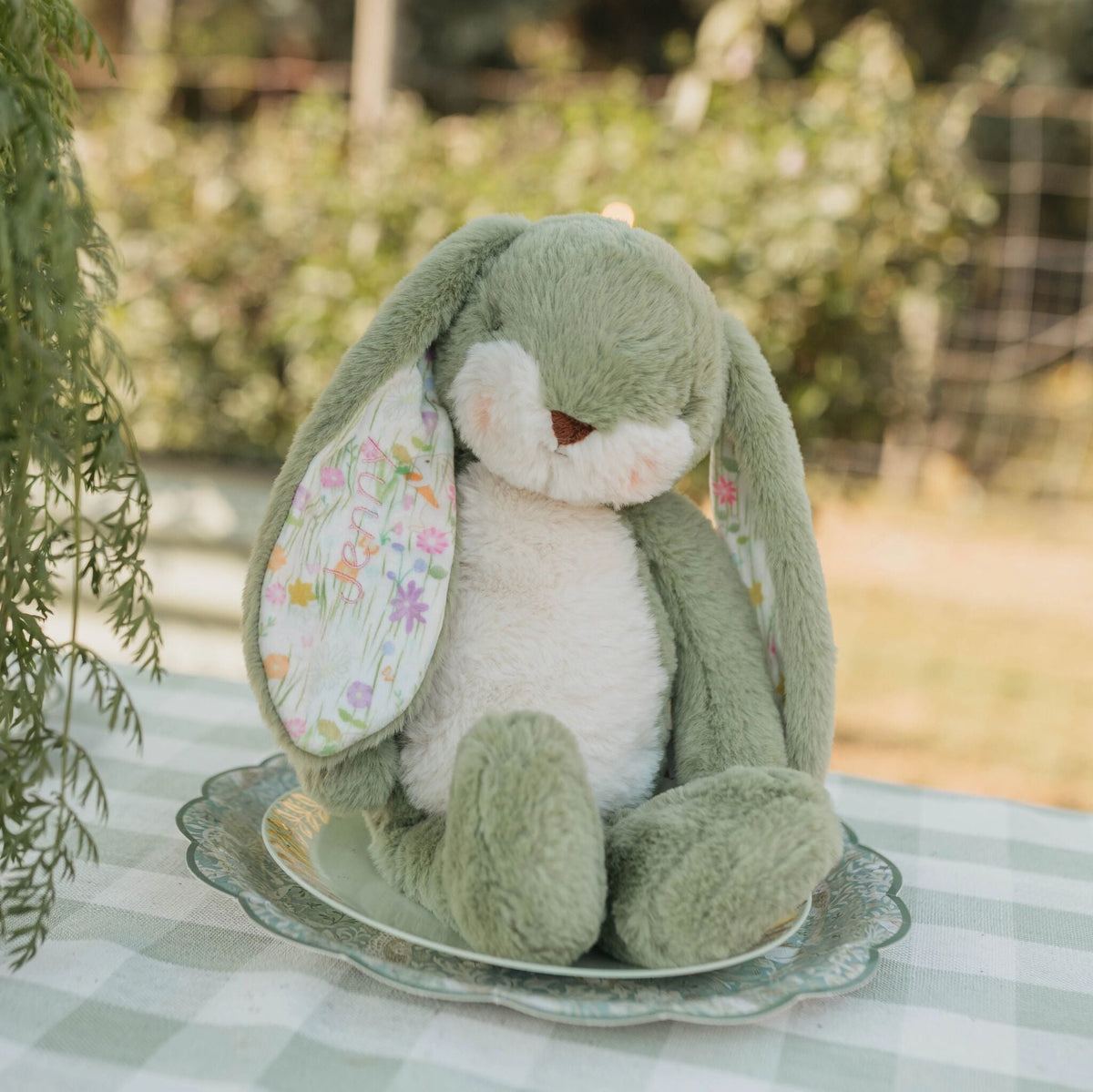 A plush bunny toy with long floppy ears sits on a decorative plate on a checkered tablecloth outdoors. The bunny is green with a white belly and face, and one ear has a floral print.