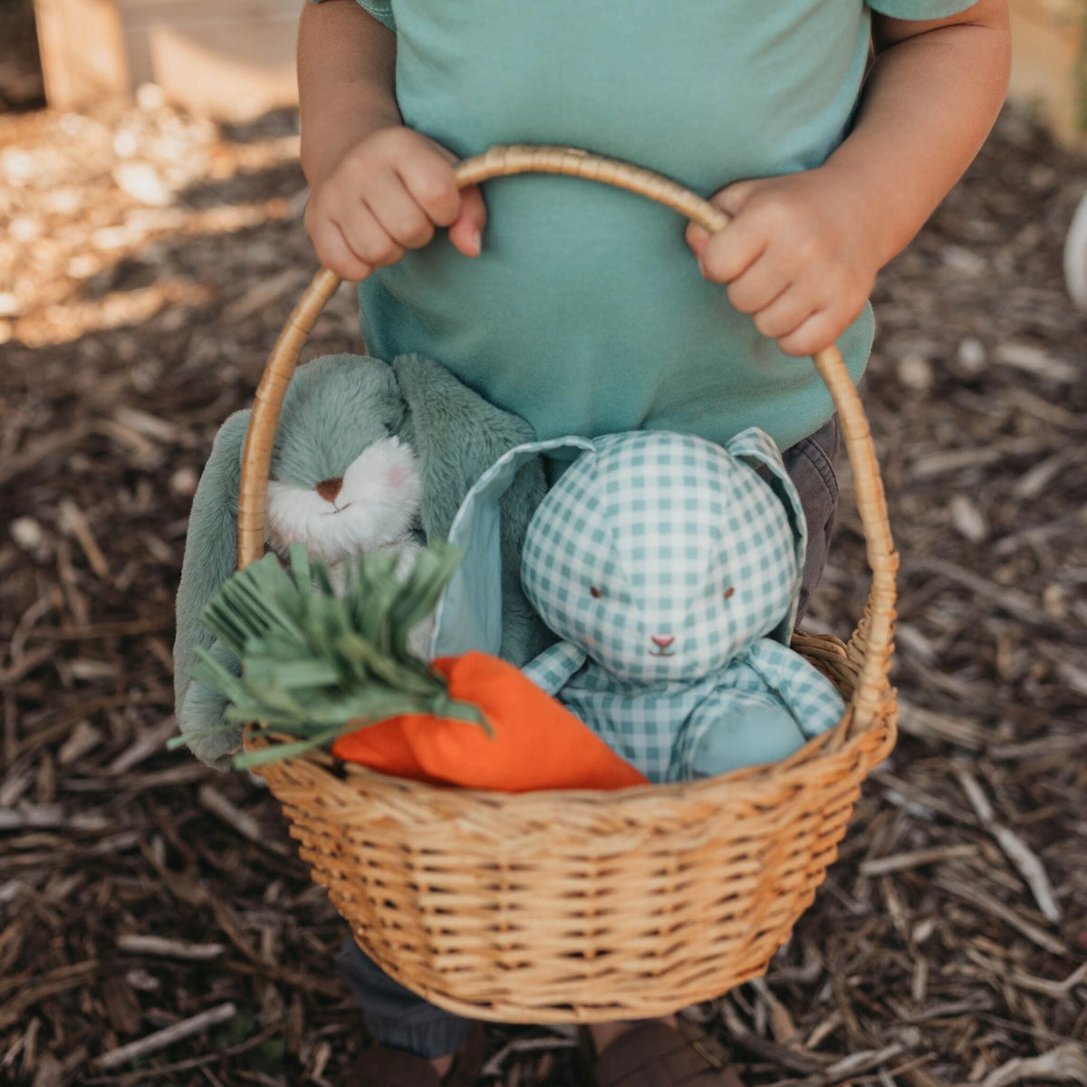 A child in a teal shirt holds a wicker basket filled with plush toys, including a blue bunny, a light blue gingham bunny, and an orange carrot, outdoors on a mulch ground.