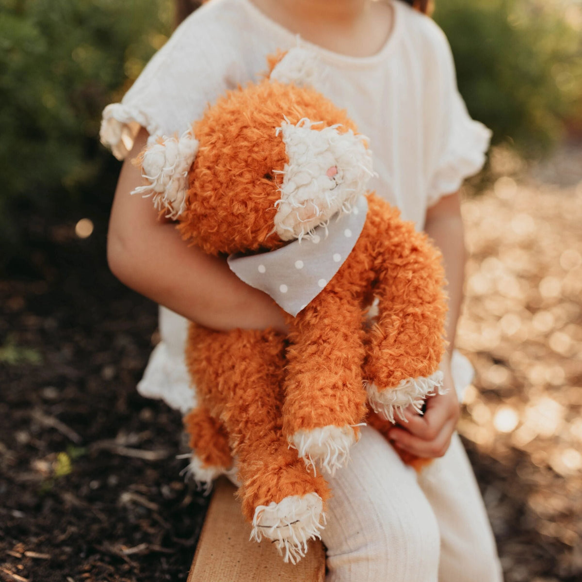 A child in a white dress holds a fluffy orange stuffed cat with white accents and a gray polka-dot bandana outdoors.