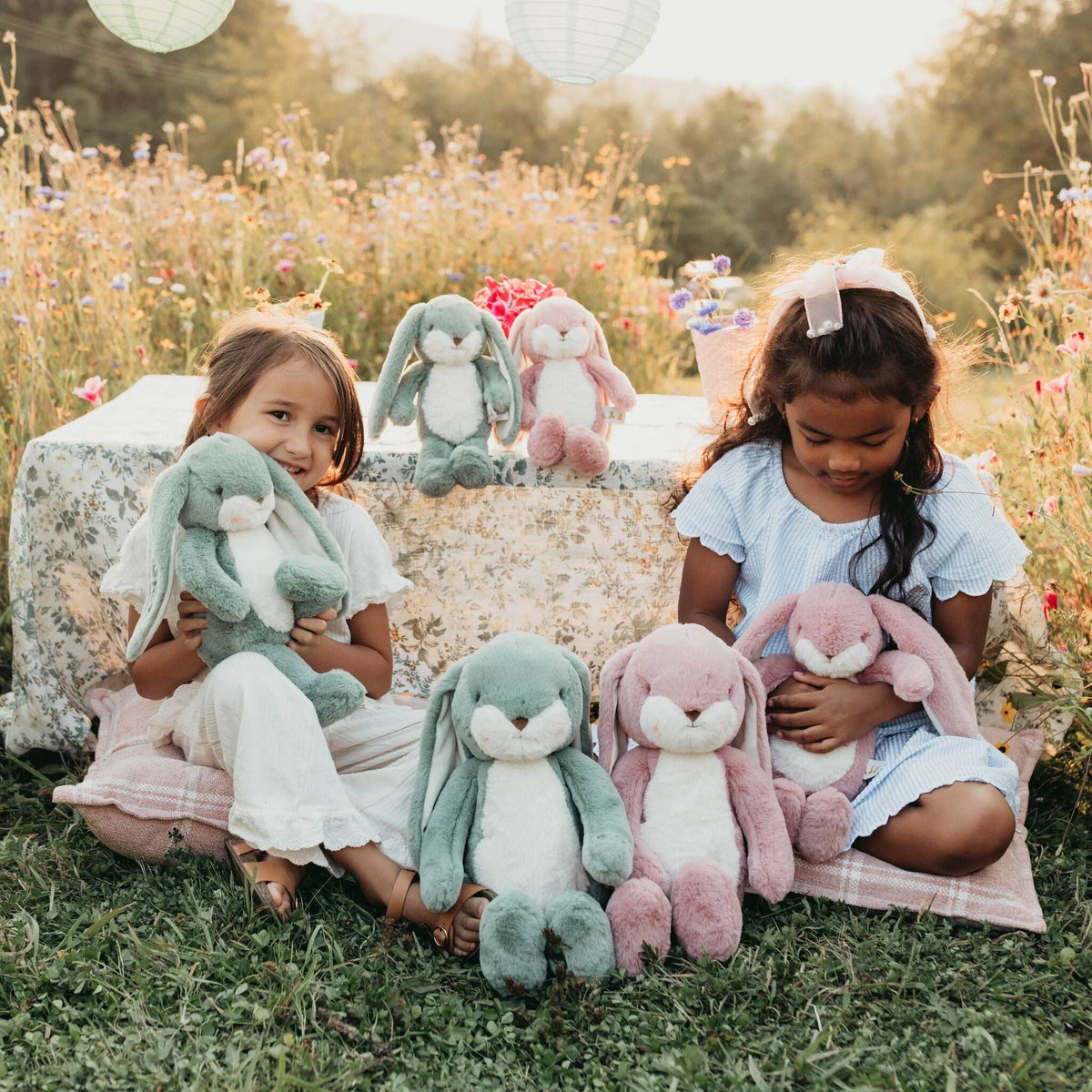 Two young girls sitting outdoors on a grassy field beside a table, each holding a plush bunny, with additional plush bunnies displayed on and beside the table. The scene is styled in a warm, sunlit meadow with flowers and lanterns in the background.