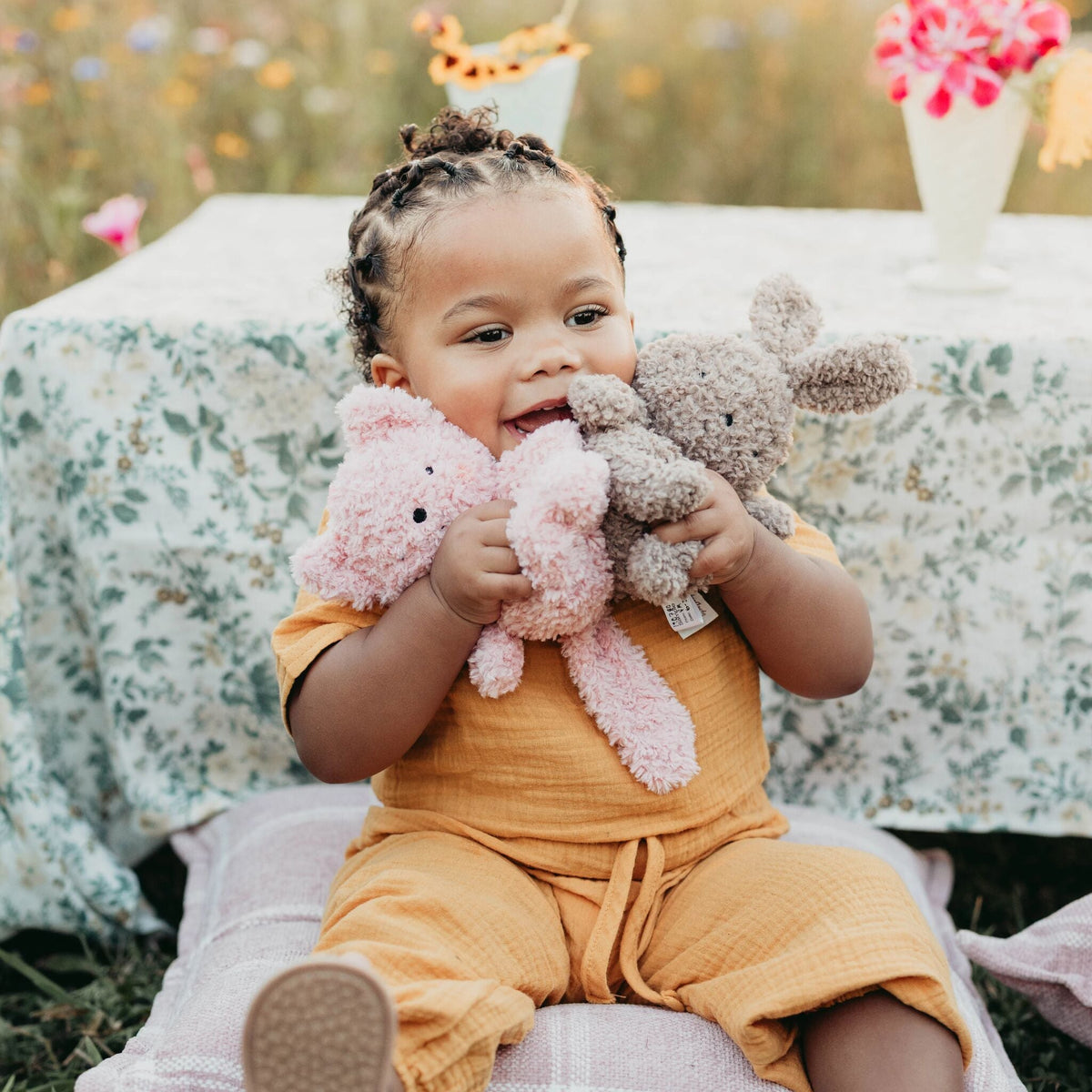 A smiling toddler sits outdoors on a blanket in front of a floral tablecloth, holding two plush stuffed animals, one pink and one taupe-gray.