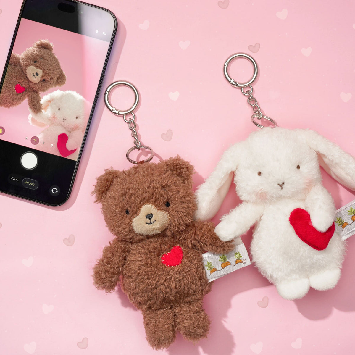 Two plush keychain figures on a pink heart-patterned background: a brown teddy bear holding a red heart and a white bunny holding a red heart. A smartphone in the corner shows a photo of the teddy bear.