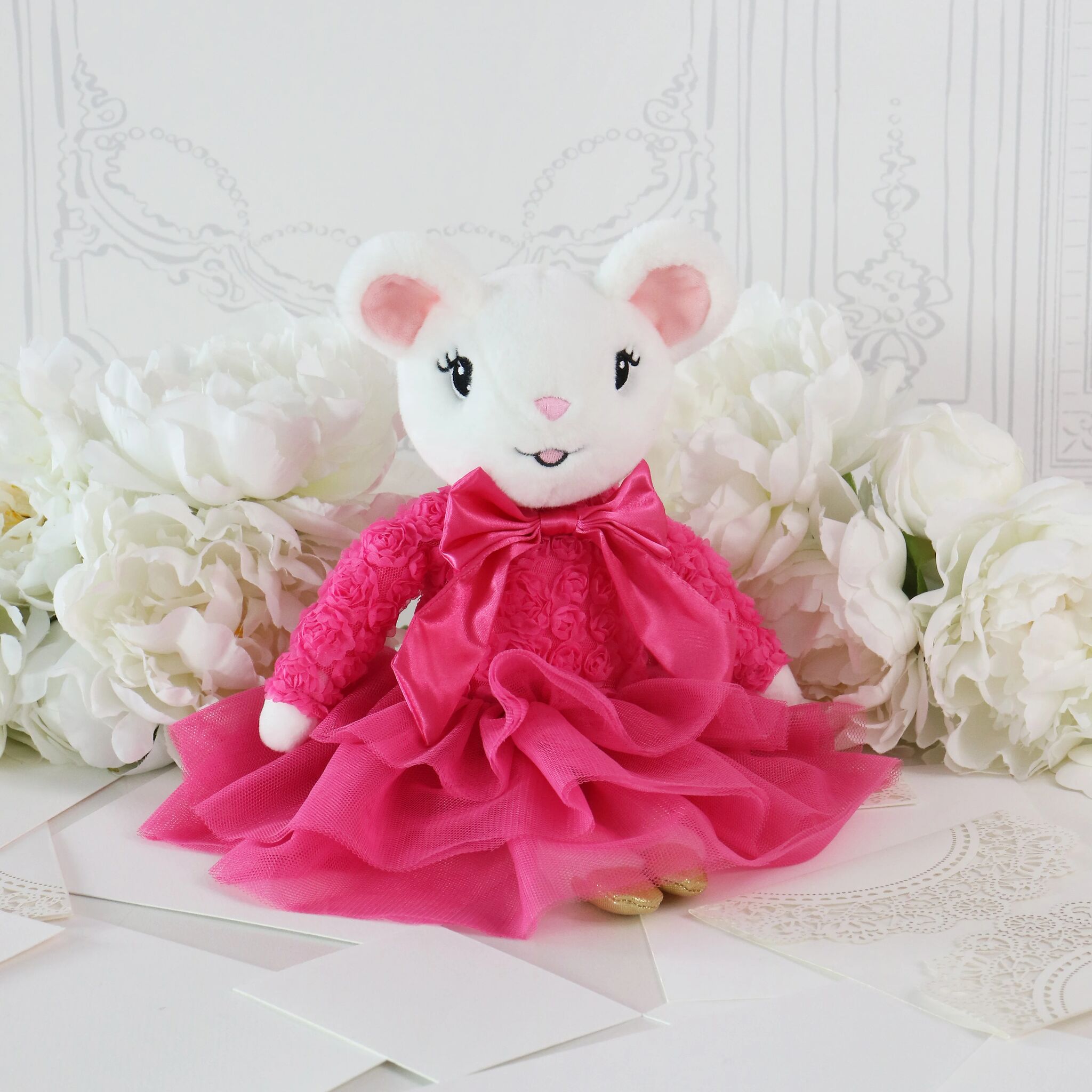 A white plush mouse toy wearing a bright pink ruffled dress and bow, posed among large white flowers on a light surface.
