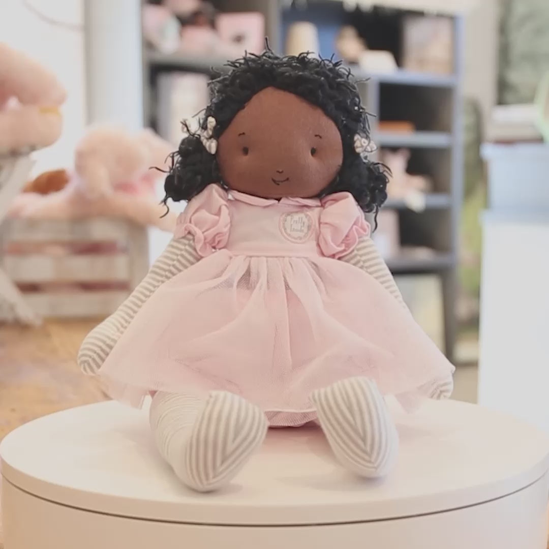 A 360 degree video of a doll with warm brown skin and black hair, in a pink dress. The doll is pictured on a white turntable with a blurred background.