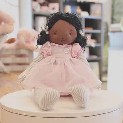 A 360 degree video of a doll with warm brown skin and black hair, in a pink dress. The doll is pictured on a white turntable with a blurred background.
